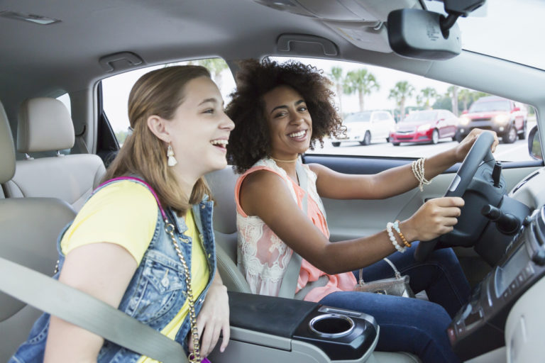 Two multi-ethnic teenage girls in a car. The focus is on the mixed race African-American and Caucasian girl in the driver's seat, smiling at the camera.