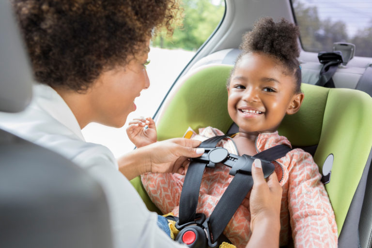 An adorable preschool age girl looks away and smiles as her unrecognizable mother buckles her into her car seat.